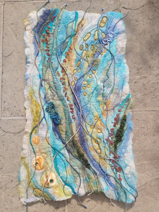 A highly textural abstract wet felted Wallhanging in shades of blue/green/gold.