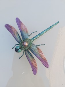 A 3D free standing fabric dragonfly made from cotton fabric, hand painted in shades of pink and turquoise. It’s wire legs have been wrapped with thread and painted to match its body.