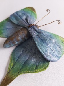 A 3D butterfly brooch made from cotton velvet, hand painted in shades of blue and green with a deep brown body.