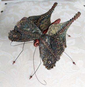 A 3D fabric butterfly constructed from cotton fabric covered with hand painted in shades of gold and dark green and stitched lace. It has long thin legs made from wire, its body is a deep russet colour