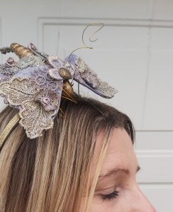 A butterfly fascinator attached to a gold coloured head band. The butterfly is made from hand painted and stitched cotton fabric and lace. It has a gold coloured body and it’s wings are a delicate mix of dusky pink and lilac